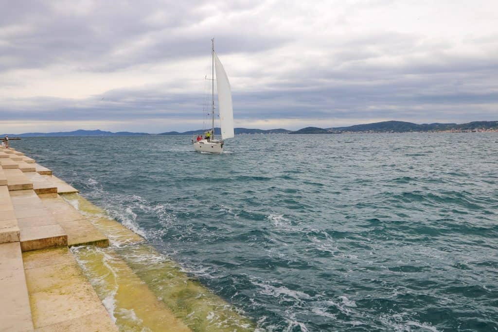 Walking along the water in Zadar Old Town looking at a sail boat passing by the steps of the Sea Organ and one of the best things to do in Zadar, Croatia. 