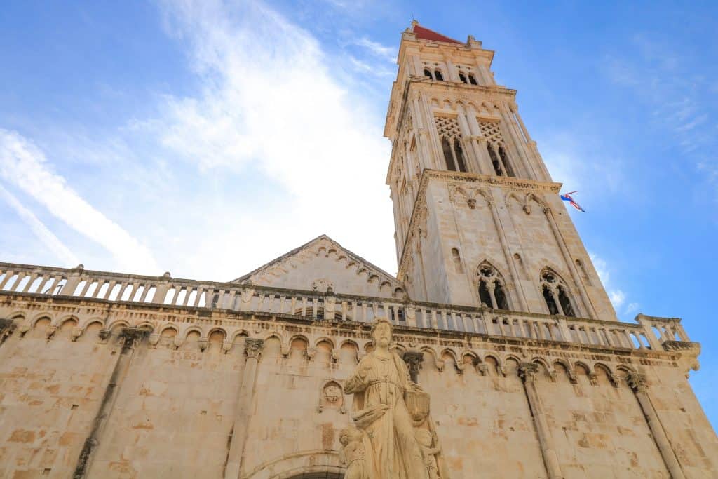 Standing at the base of St. Lawrence Cathedral & bell tower looking up at its magnificent architecture with a statue in front on a sunny day in Trogir.