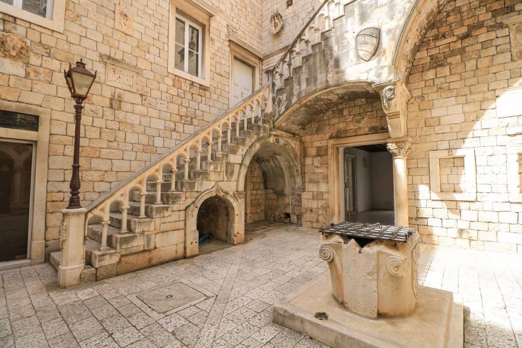 The beautiful courtyard of the Town Hall all made of marble and bricks in white and tan colors with a ornate staircase, tall lantern and a well in the center of it in Trogir, Croatia.