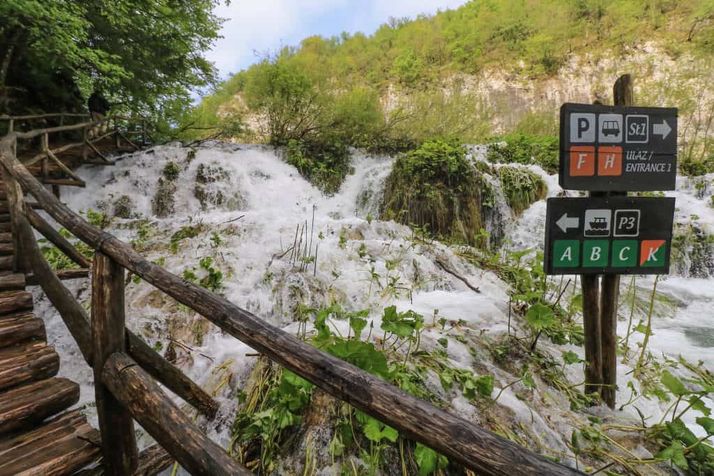 Heavy flow of water cascading down the rocks and into the lower lake from a boardwalk with railing and one of many helpful signs with route info on it at Plitvice Lakes.