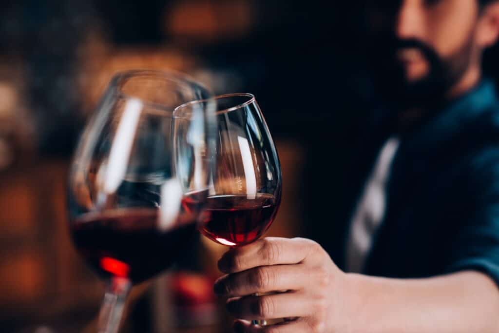 A man holding a glass of red wine up to another glass of wine another person is holding during a wine tasting.