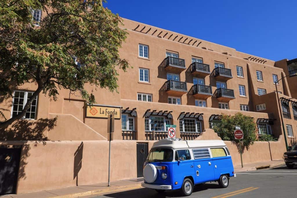 The tan pueblo style of architecture of the historic La Fonda Hotel was also once a "Harvey House" with a bright blue VW bus parked in front in Santa Fe, NM.