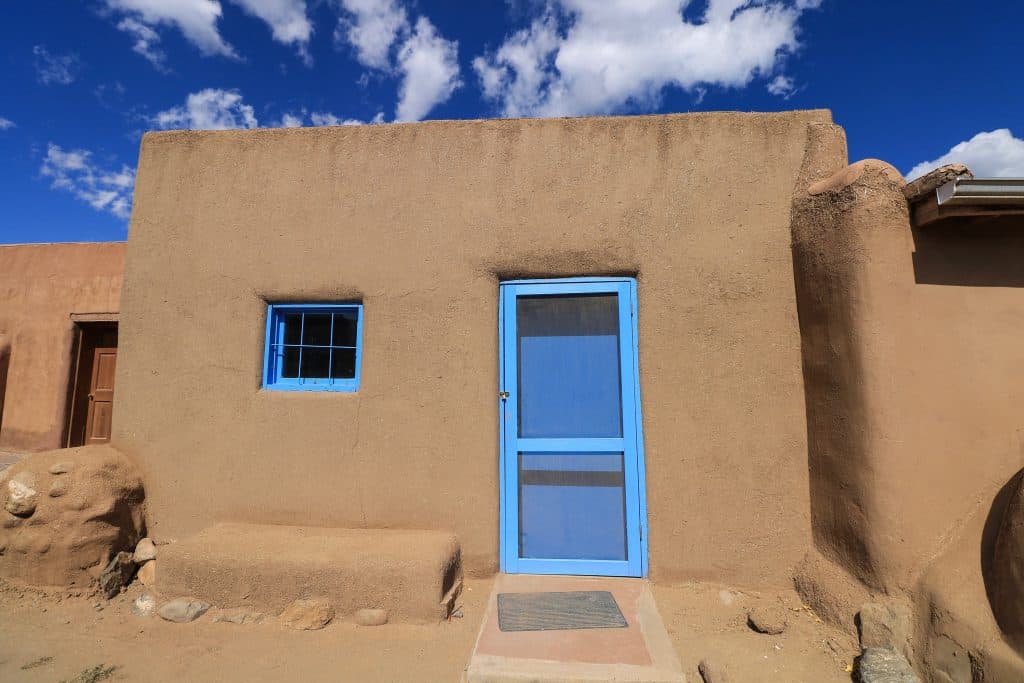 A turquoise door and window on a tan and small adobe house where someone lives in Taos Pueblo, a World Heritage Site.