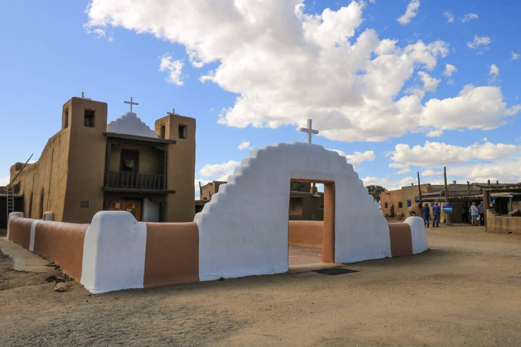 The beautiful chapel at Taos Pueblo in tan adobe style and a white and tan wall surrounding it are where tours start every 20 minutes on a sunny day with puffy clouds.