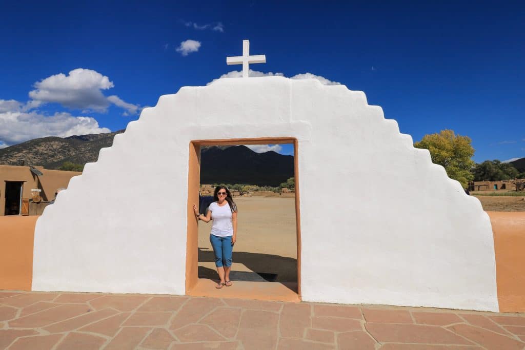 Standing at the entrance to the chapel at Taos Pueblo underneath the white arched doorway to the courtyard with a cross on top of it.
