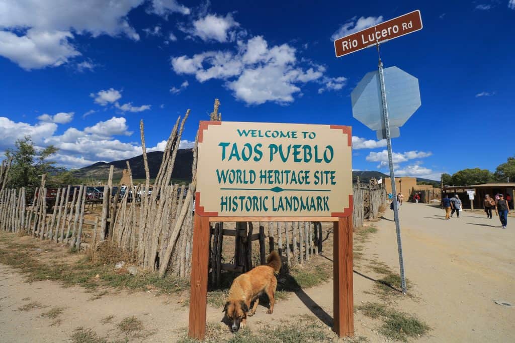 Taos Pueblo entrance with a large sign against a fence and a tan dog in front in the town of Taos, New Mexico.