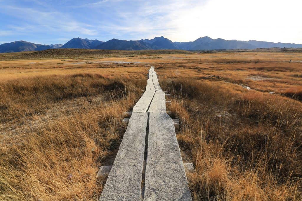 A thin wooden boardwalk leading out through a flat landscape to a natural hot spring with mountains in the distance near Mammoth Lakes, CA.