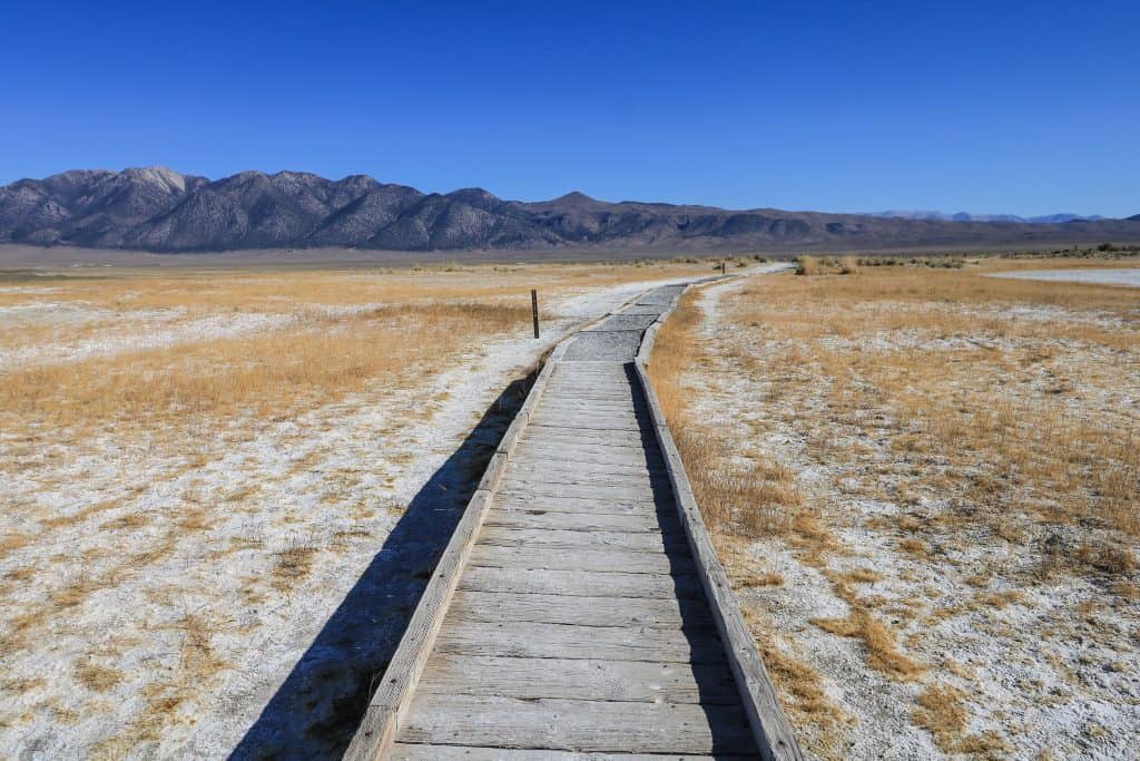 Walking on the boardwalk path to Wild Willy's hot springs on a flat plain area and tall mountains in the distance in Mammoth Lakes, CA.