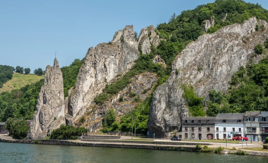 View of Rocher Bayard rock along the Meuse River surrounded by green foliage from across on the other side of the river in Dinant, Belgium.