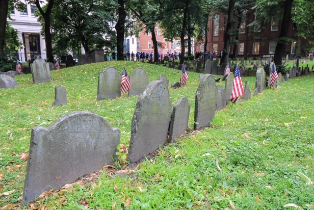Rows of short and old headstone markers with small American Flags stuck in the grass in between at the Granary Burying Ground in Boston.