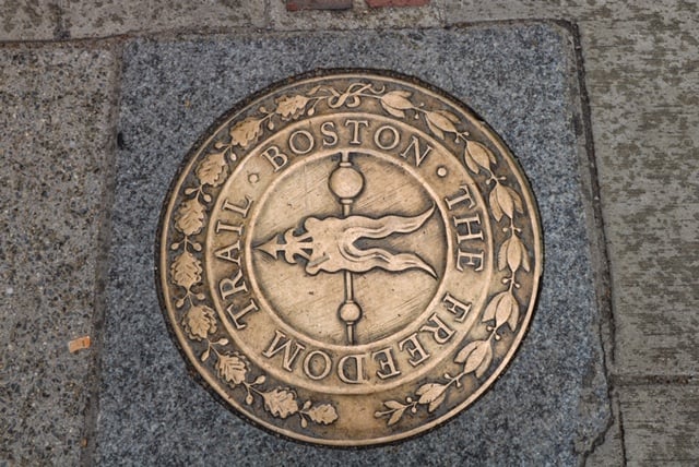 A round ornate metal plaque on the ground in Boston Common has the wording The Boston Freedom Trail as a marker to where it begins.