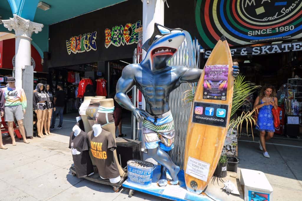 A fun shop on the boardwalk with a surfer shark statue who is extremely buff in front of a surf shop