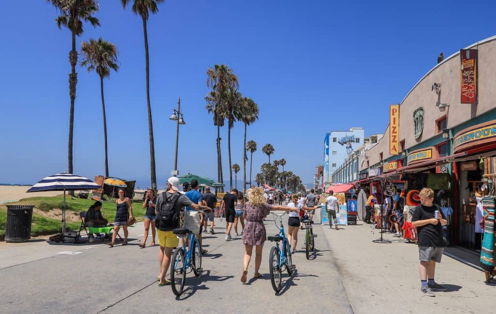 People walking and riding their bikes along the Venice Beach boardwalk with palm trees lining the way
