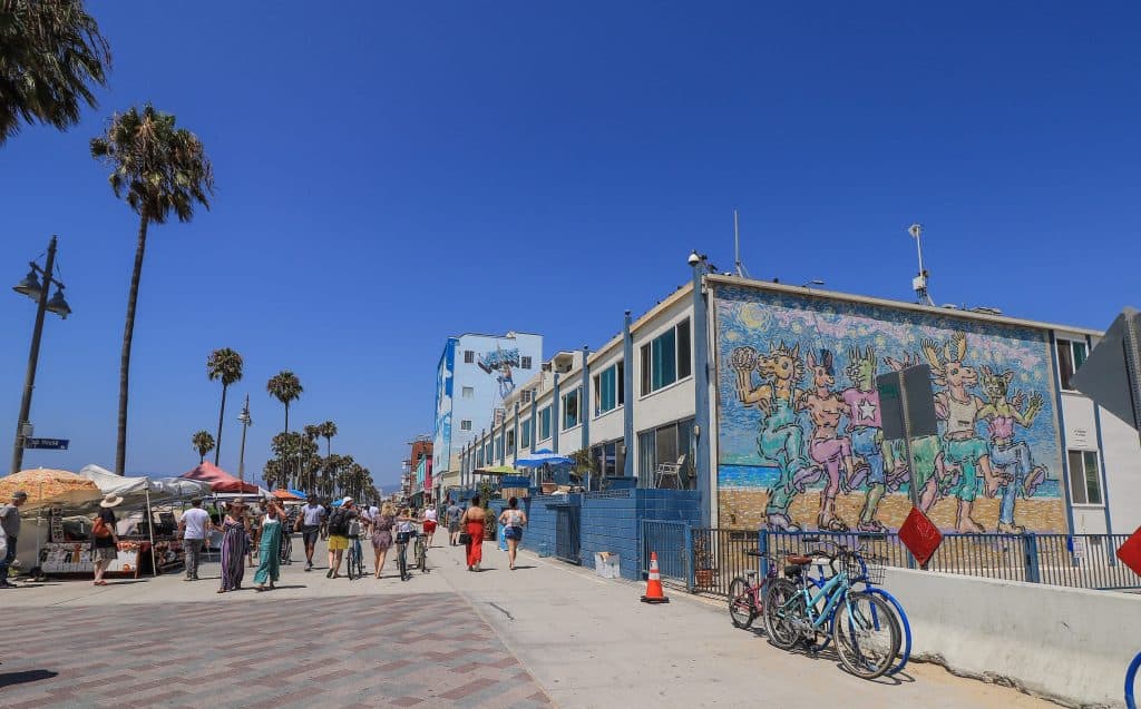 People hanging out and walking along the Venice Beach boardwalk and with a huge fun art mural on the side of one building.