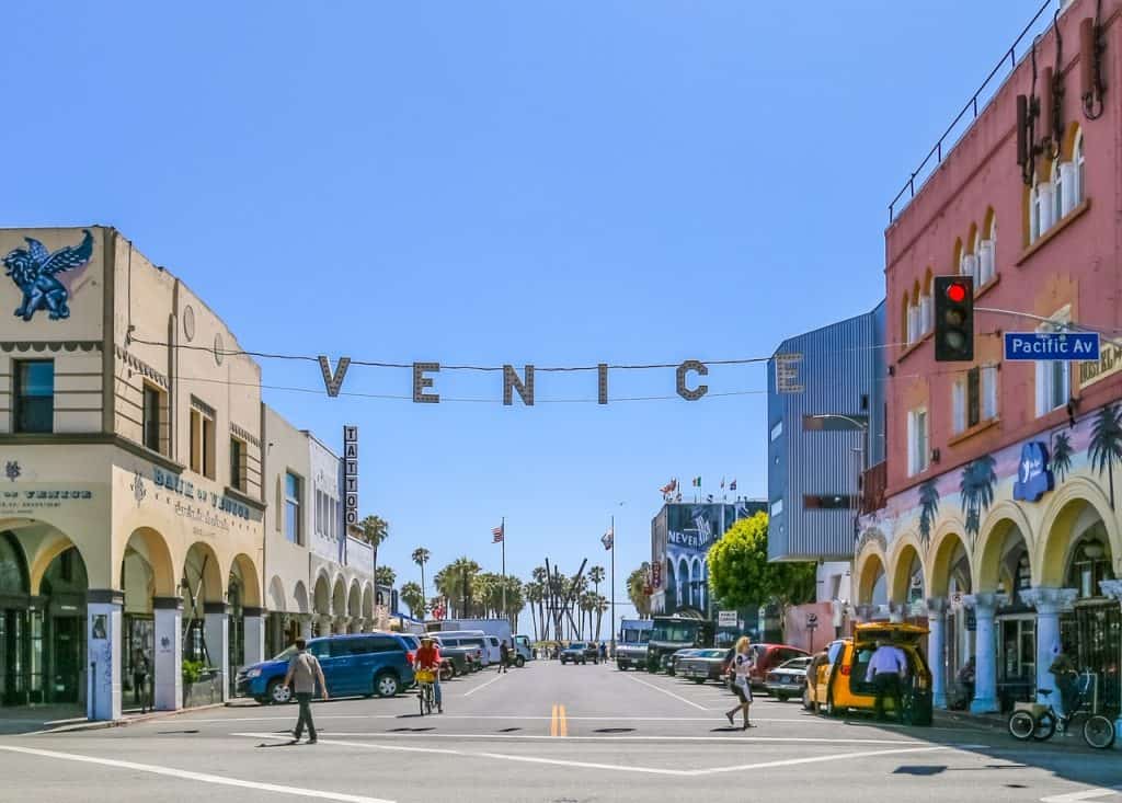 Venice Beach, USA - The Venice sign hanging over Windward Avenue at daytime. There are signs affixed to houses saying tattoo, hotel and hostel. People are walking by and many cars are parked. Mural paintings are also part of the scenery, as well as the beach and the pacific ocean in the background.