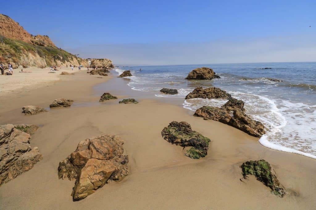 A gorgeous section of a Malibu beach with golden sand and a number of small to medium sized rocks in the shoreline.