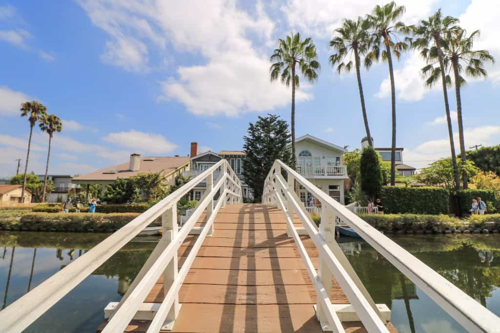Walking over a wooden bridge that is white and crosses a canal. Palm trees are across the way with a very nice house.