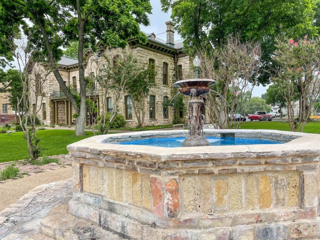 A stone water fountain surrounded by grass and a German-styled building in the backdrop in Fredericksburg.