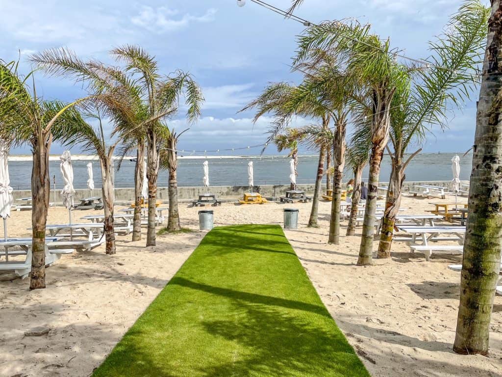 A long fake grass carpet lined with palm trees leading out to tables near the beach edge at The Gulf in Orange Beach, Alabama.