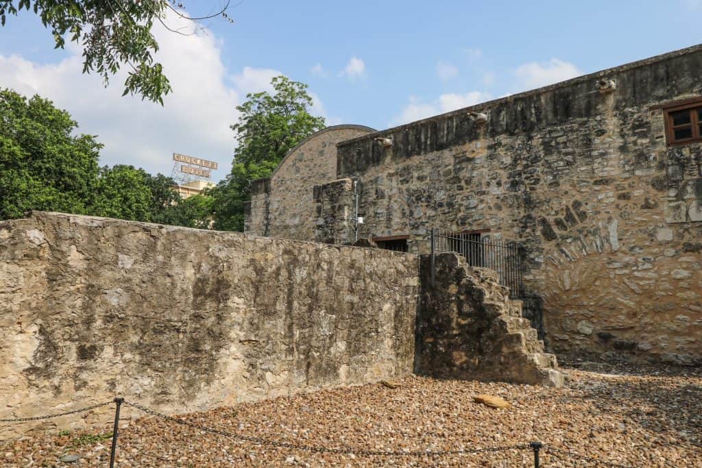 View of the side of the Alamo with its walls and in the distance a sign for the Crockett Hotel.