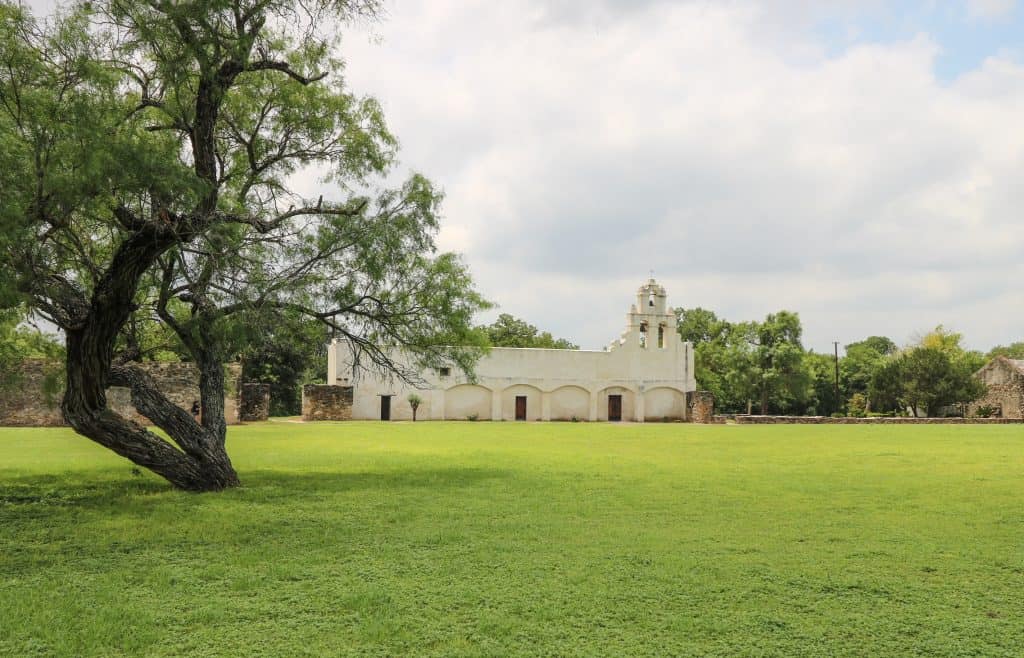 Mission San Juan stands out from the rest as it is stark white with bells at the top surrounded be a large green lawn and trees.