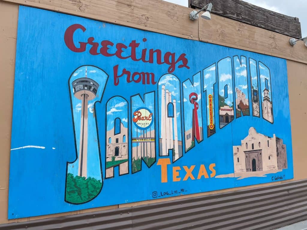 The older Greetings from San Antonio art mural that is blue with scenes of San Antonio painted on each letter.
