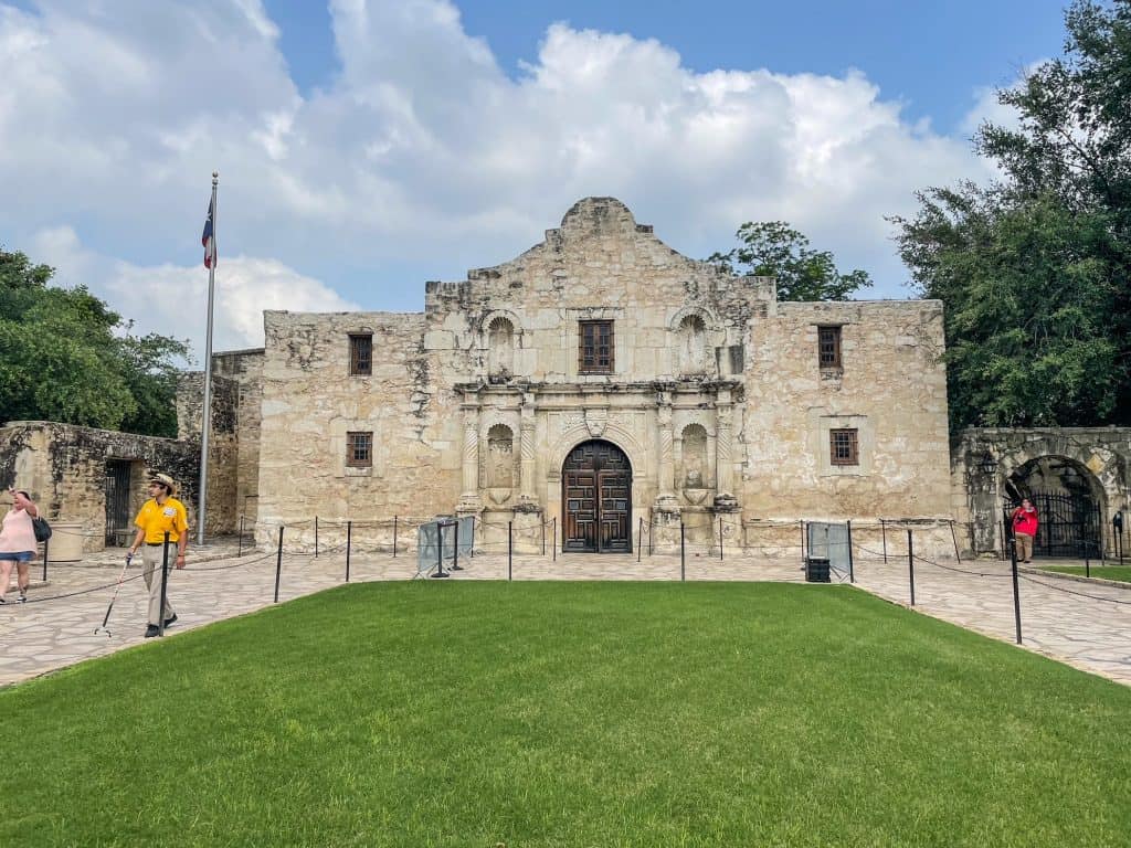 The famous Alamo made of limestone, a green patch of grass in front and a Texas flag blowing in the wind.