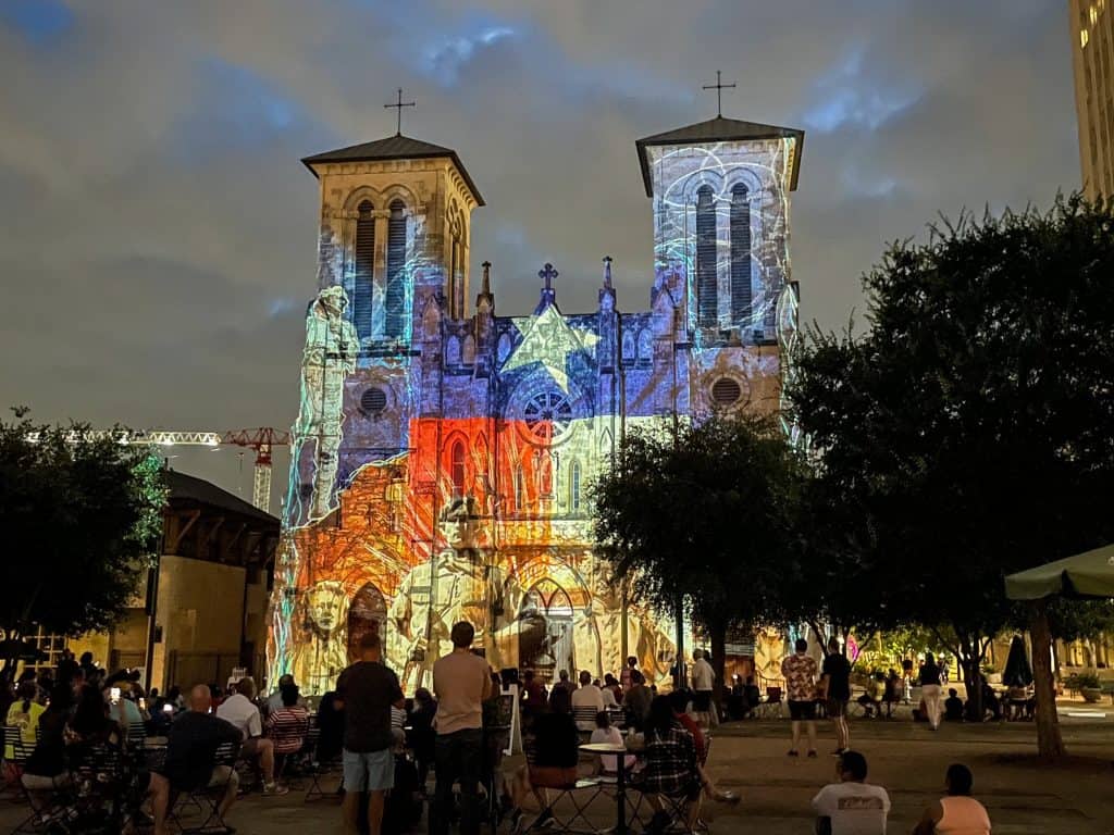 San Fernando Cathedral lit up at night with bright colors of the Saga Light Show displaying on the front with the Texas flag.