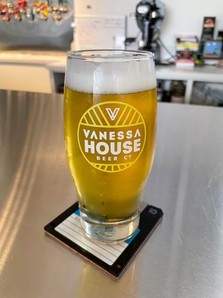 A cold beer in a glass that has the Vanessa House Beer logo on a floppy disk coaster.