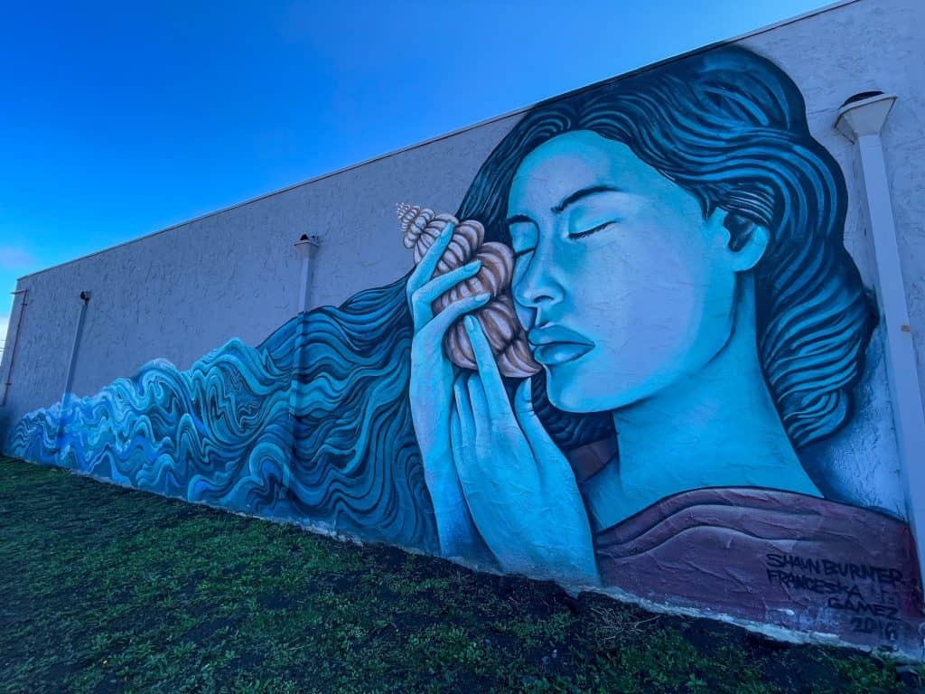 A beautiful art mural in shades of blue of a woman holding a sea shell to her cheek and flowing hair the ocean.