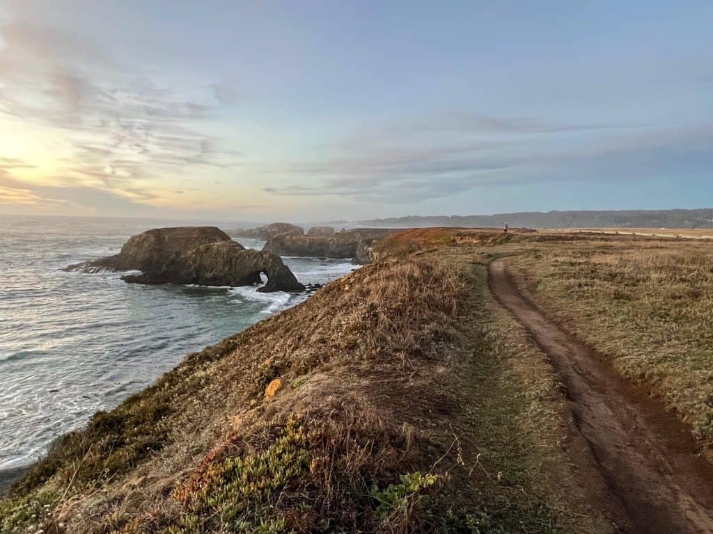 Walking along a thin trail looking out at the ocean and a sea arch at sunset.