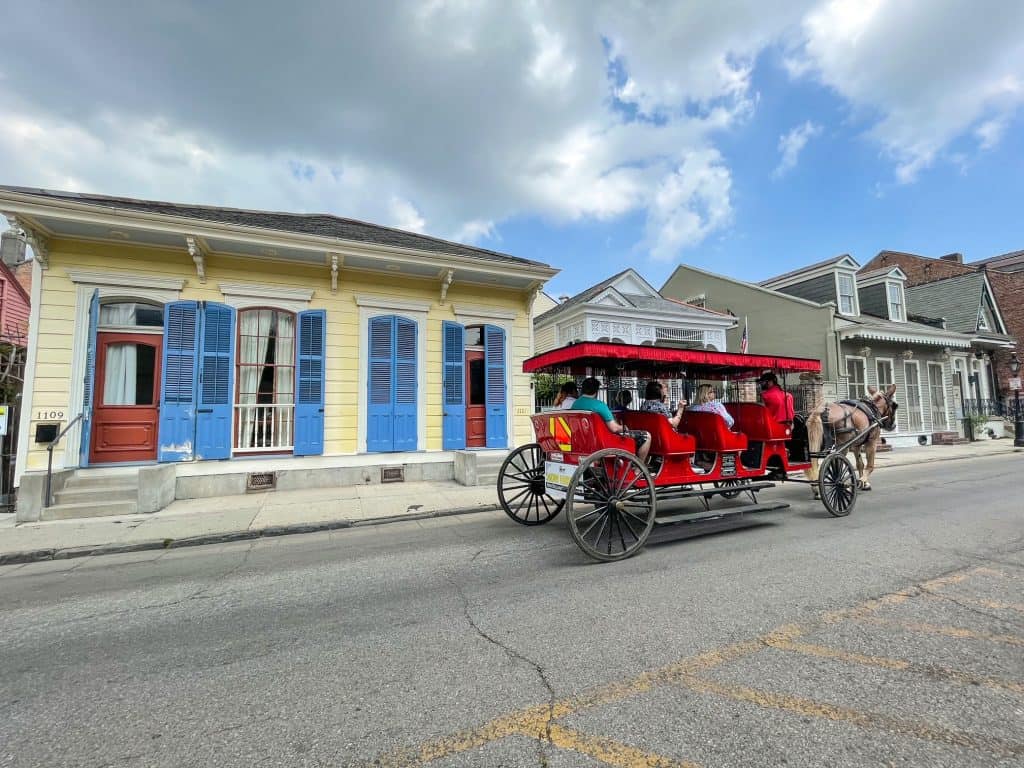 A horse drawn red carriage passing by a yellow house with blue shutters in New Orleans.