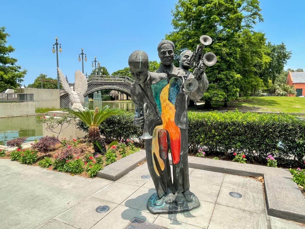 A sculpture of three men playing instruments as one body with a pond, bridge, and flowers in the background.