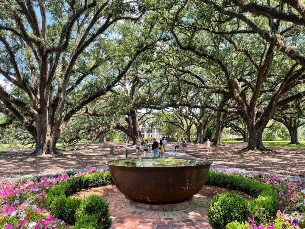 A huge copper bowl filled with flowers and a brick flooring with the mansion and row of oak trees behind.