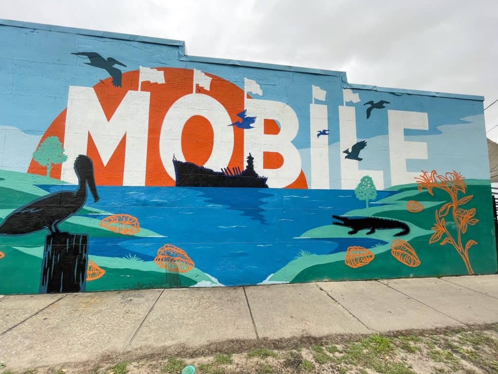 The Mobile art mural with letters in white against a blue, green and orange scene of the sun over Mobile Bay is one of many murals to see and one of the fun things to do in Mobile, Alabama.