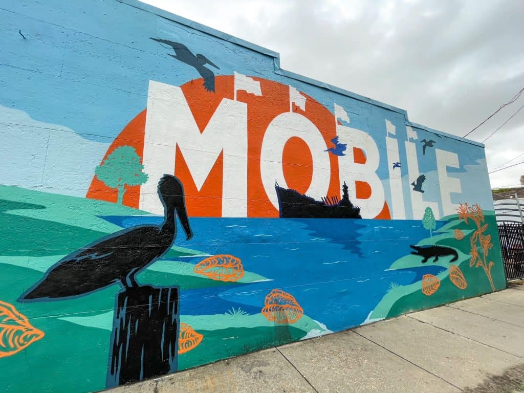 A colorful mural with Mobile written on it and scene of Mobile Bay with pelican and the USS Alabama.