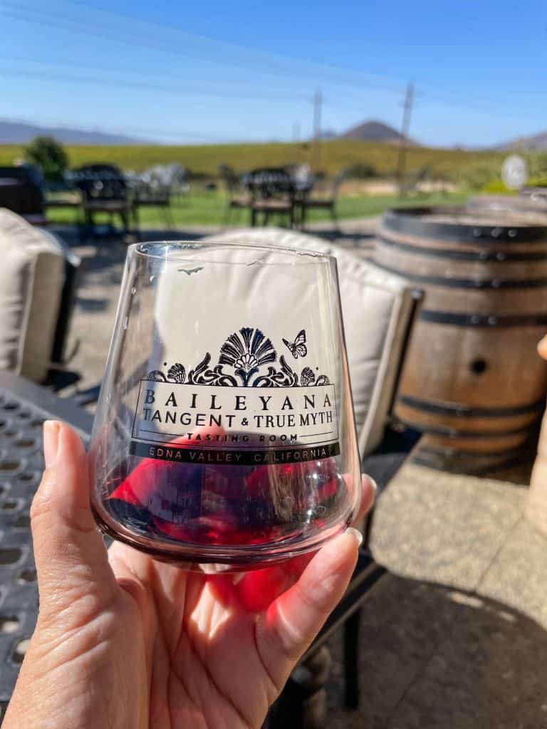 Holding a glass of Pinot Noir with a wine barrel and Islay Hill in the background at Baileyana Vineyard in San Luis Obispo, California.