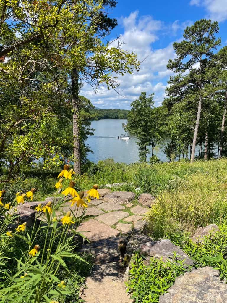 Lake Hamilton between the trees with a boat going by and wildflowers, grass, and stone pavers at Perry Wildflower Overlook at Garvan Woodland Gardens in Hot Springs, AR.