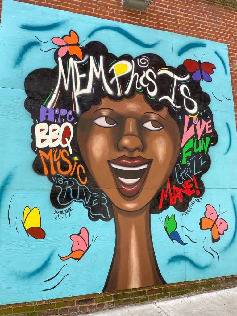An art mural with a bright turquoise background with a black woman and words describing Memphis are written in her hair.