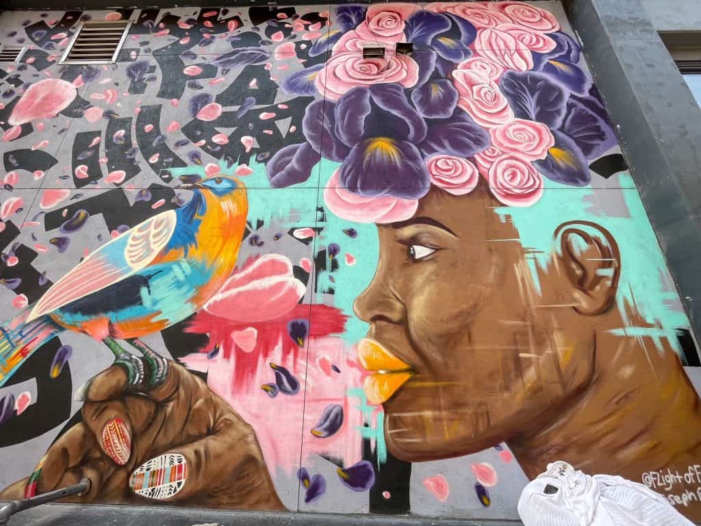 A mural with a black woman painted with her hair purple an pink flowers, flower petals flowing everywhere and a bird on her finger.