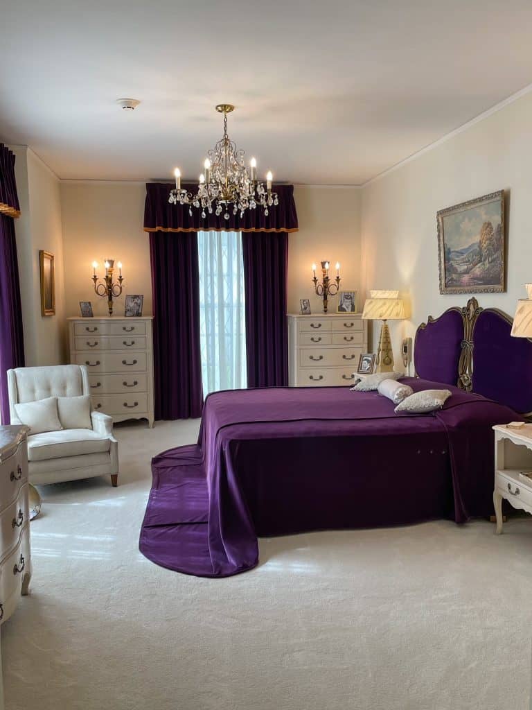 A beautiful all white carpet, chair, and dressors in a bedroom with a dark purple bedspread and curtains with a chandelier overhead in the room that belonged to Elvis's mother at the mansion.