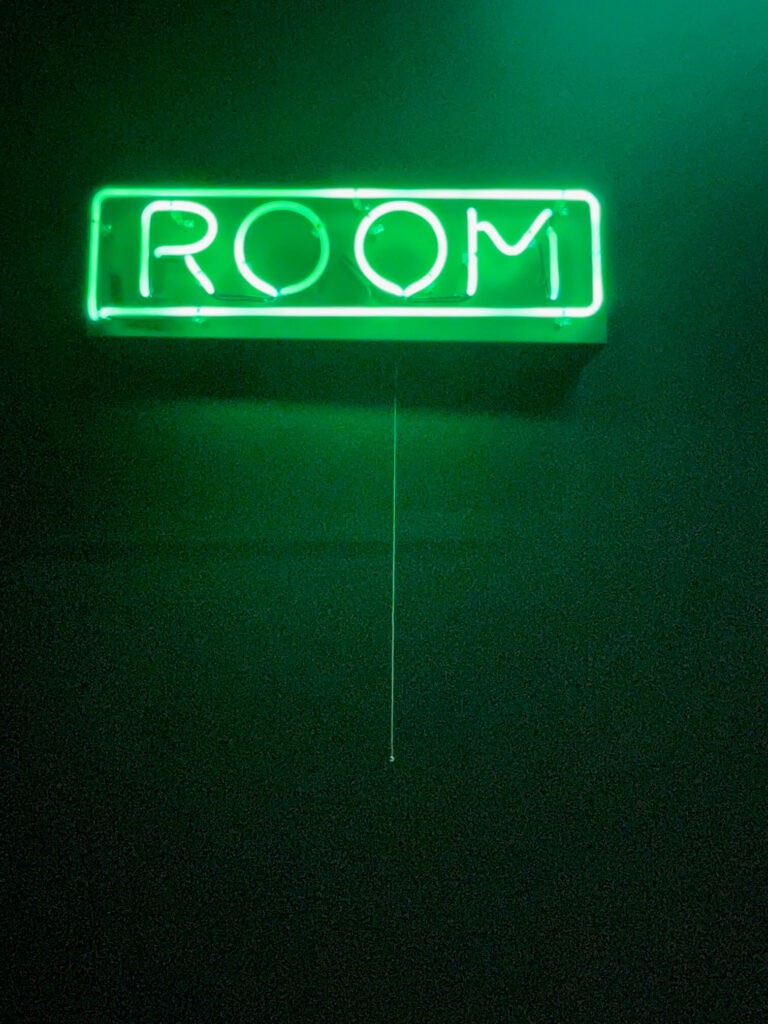 A neon green sign that says room against a dark green wall for the Green Room space.