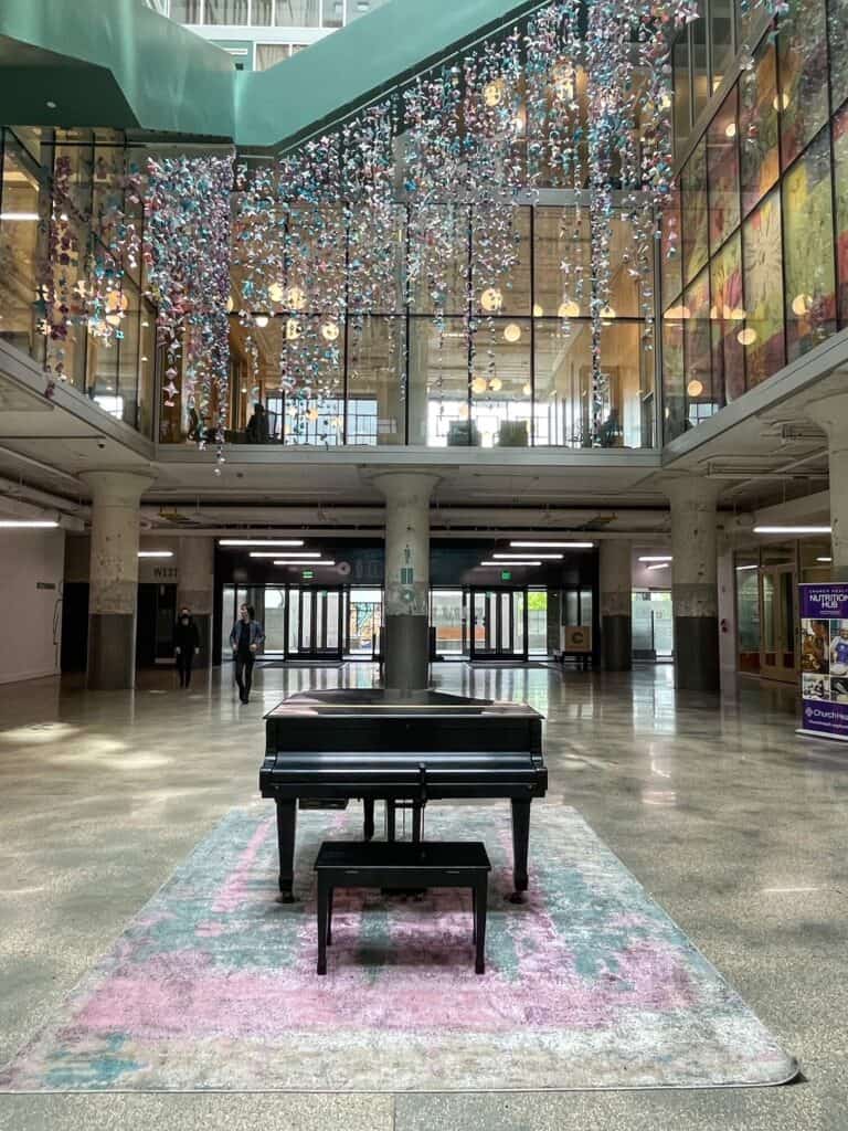 A piano on a pale pink and seafoam green rug with a beautiful art installation in the same colors hanging above resembling a chandelier.