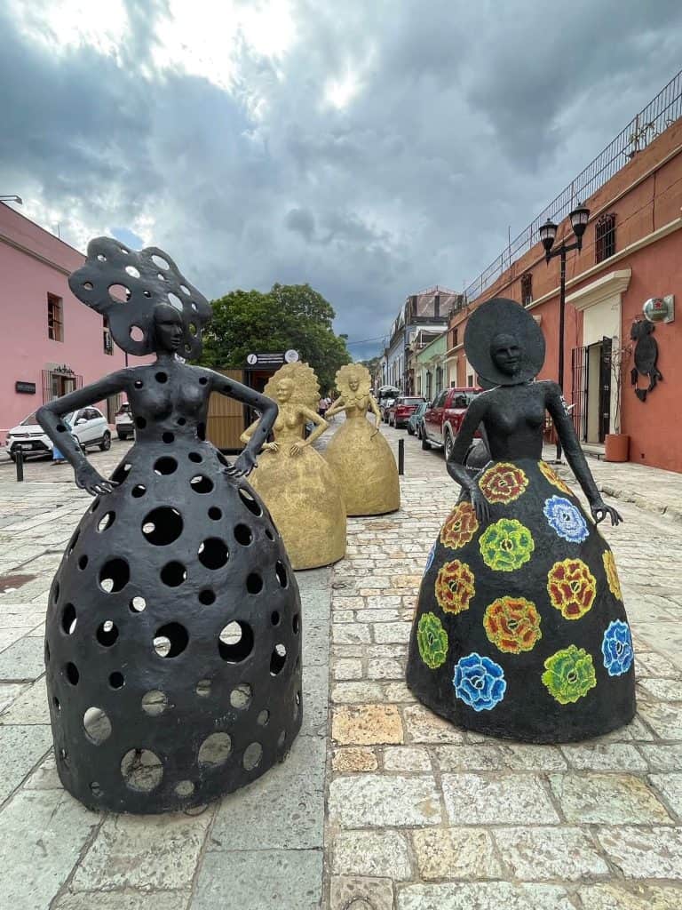 Statues on a cobblestoned street in Oaxaca City, two black and two in gold colors of females with large skirts.
