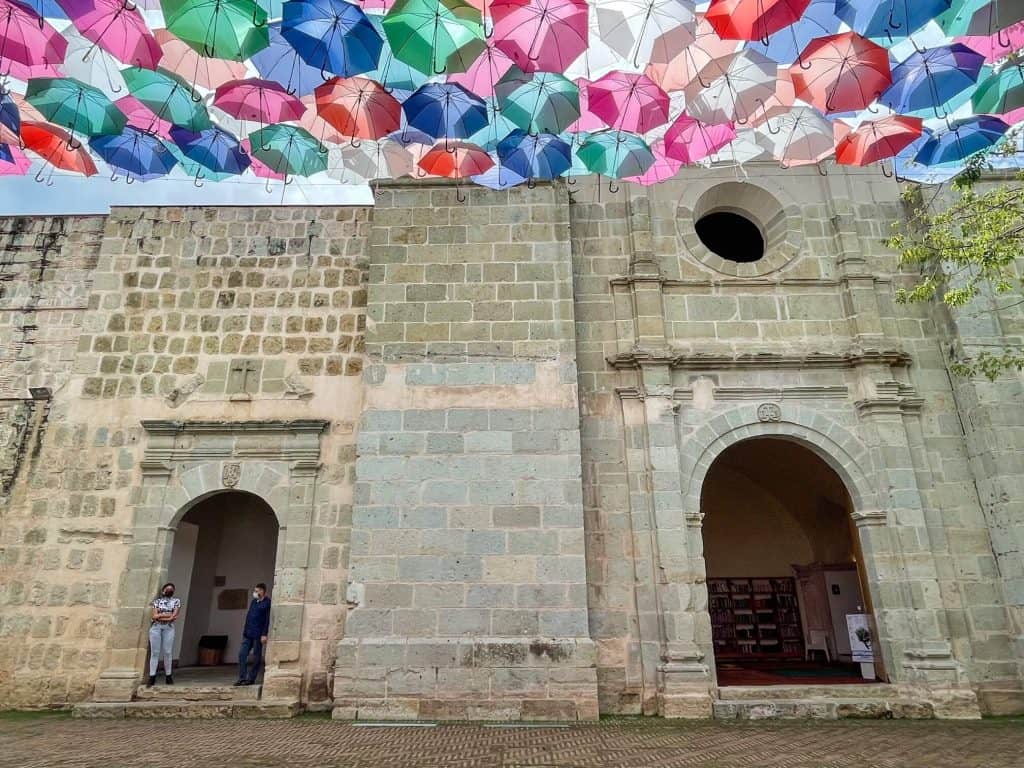 A beautiful old convent made of stone and a canopy of colorful umbrellas hanging overhead in Oaxaca.