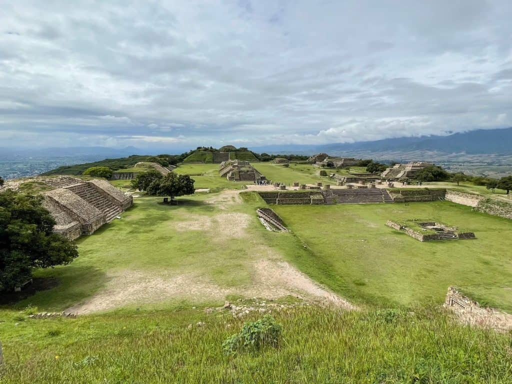 Looking down at the main valley area of Monte Alban with several pyramids and stone structures among a field of green grass.