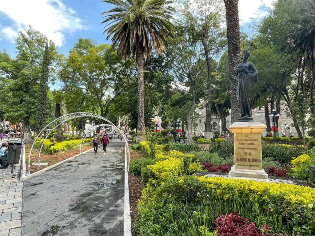 A beautiful lush park with trees. flowers, bushes, statues, and benches in the center of the Zocalo in Puebla, Mexico.