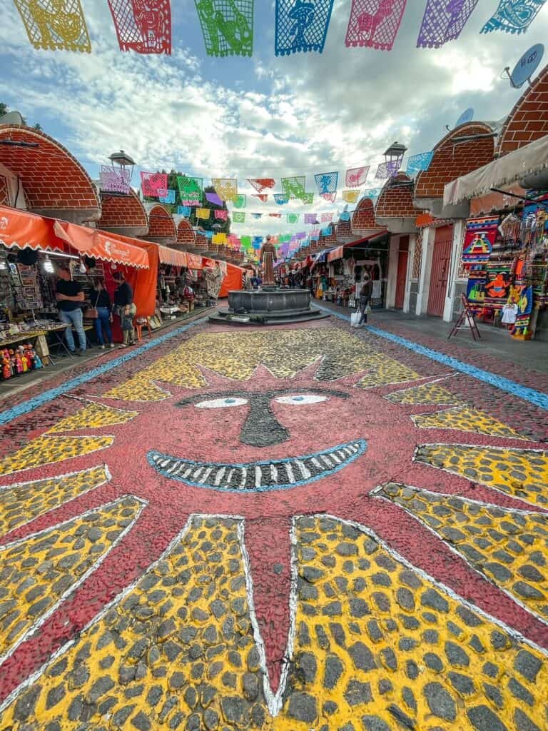 Standing in front of a huge red sun with yellow background painted on the ground at the El Parian market with vendors lining each side and flags hanging.