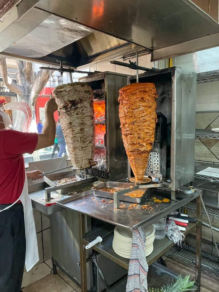 Both Arabe pork and Al Pastor pork on separate fire spits and a worker carving off the meat.