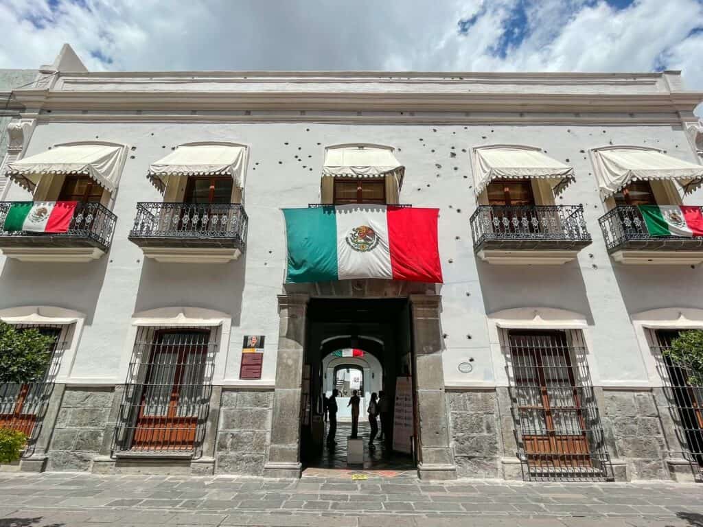 The front view of the white historic home that is now the Museum of the Revolution with several bullet holes in the facade in Puebla's historic center.